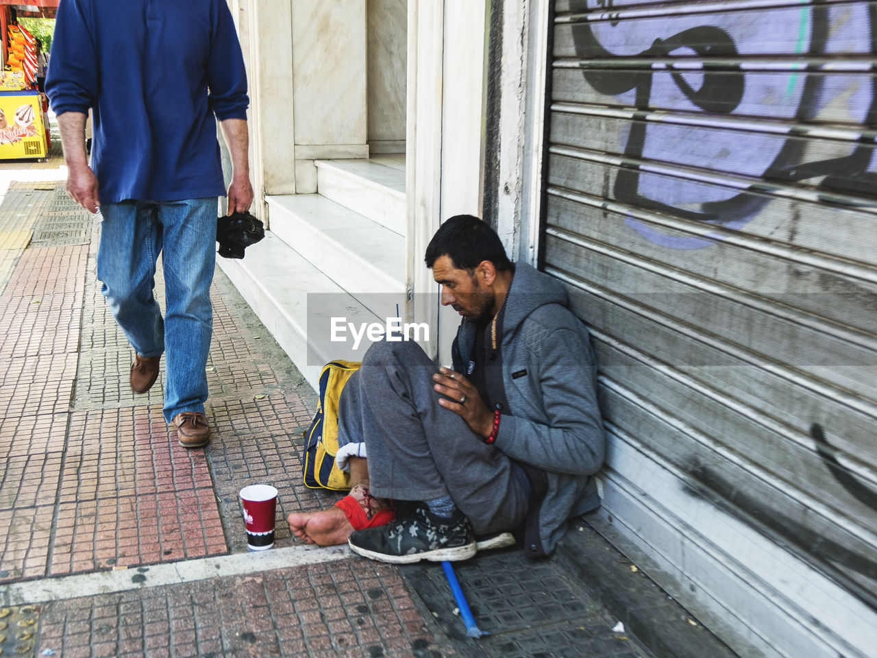 Side view of homeless man sitting in sidewalk by closed shutter