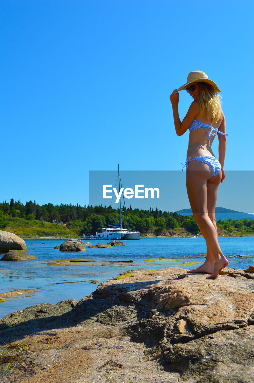 Side view of woman wearing bikini standing by sea against clear blue sky