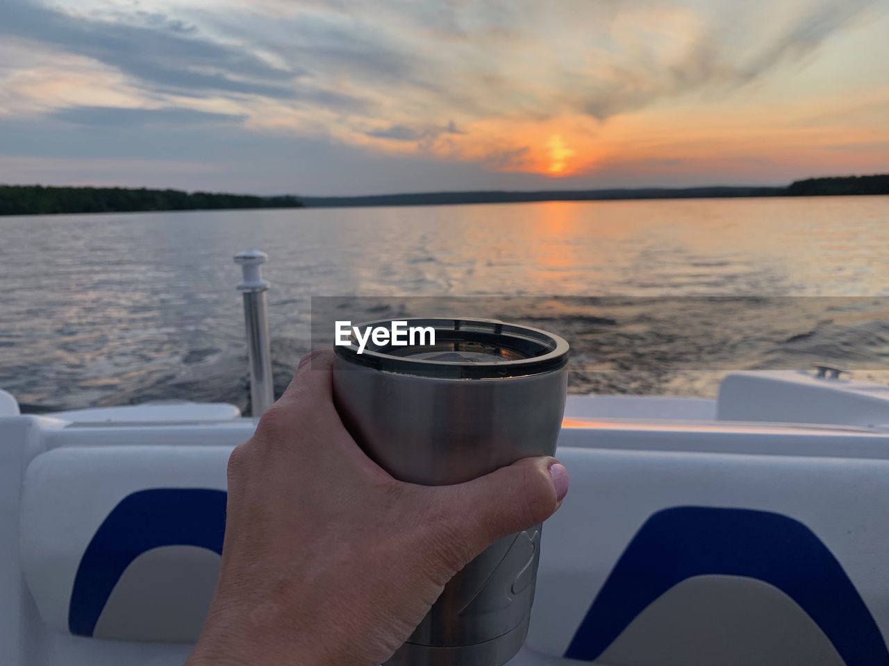 CLOSE-UP OF HAND HOLDING GLASS OF WATER AGAINST SUNSET