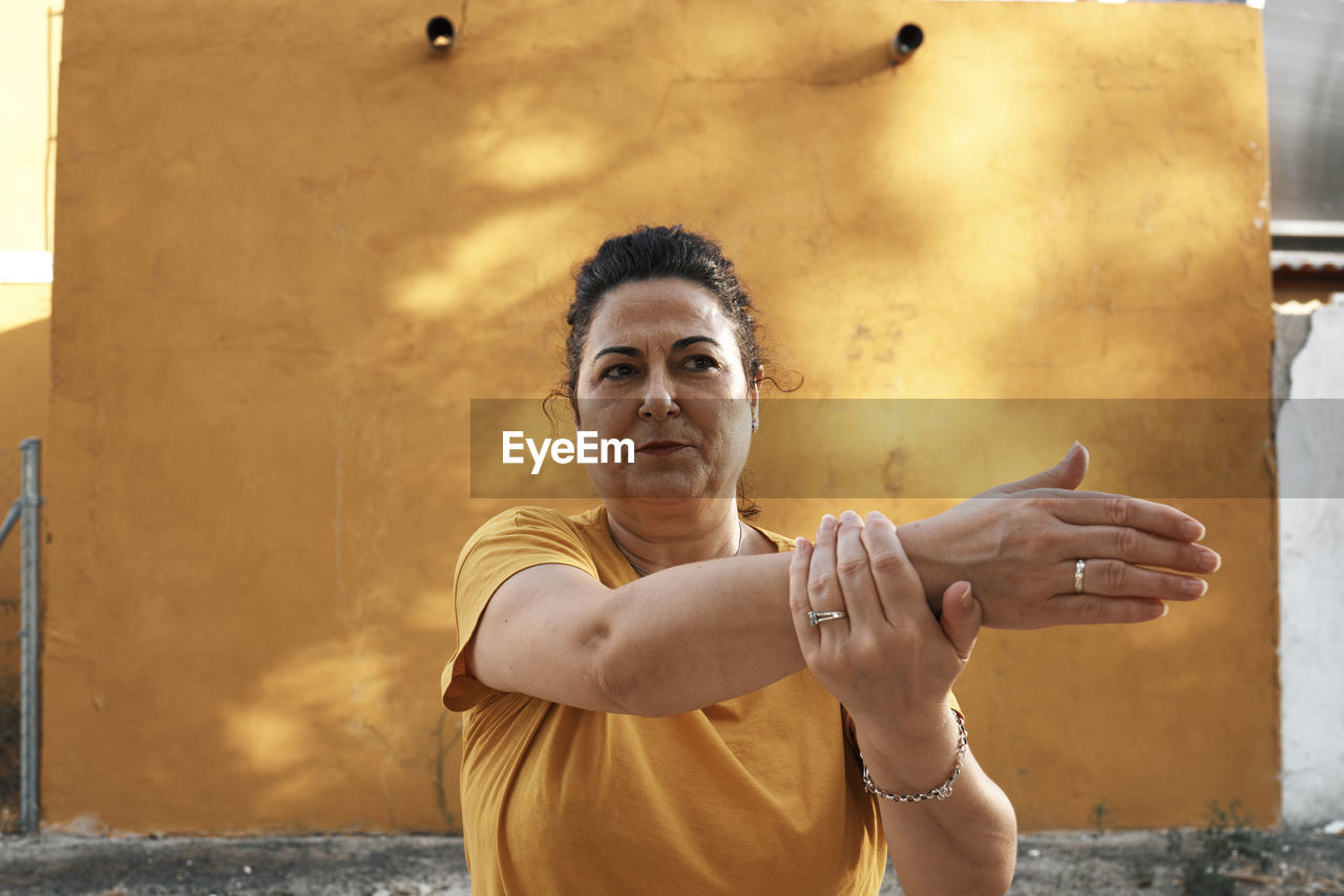 A middle-aged woman is stretching with a yellow wall in the background