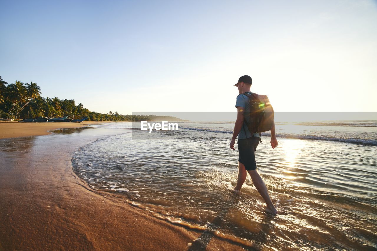 Solo traveler walking on empty beach. rear view of man with backpack at sunrise. 