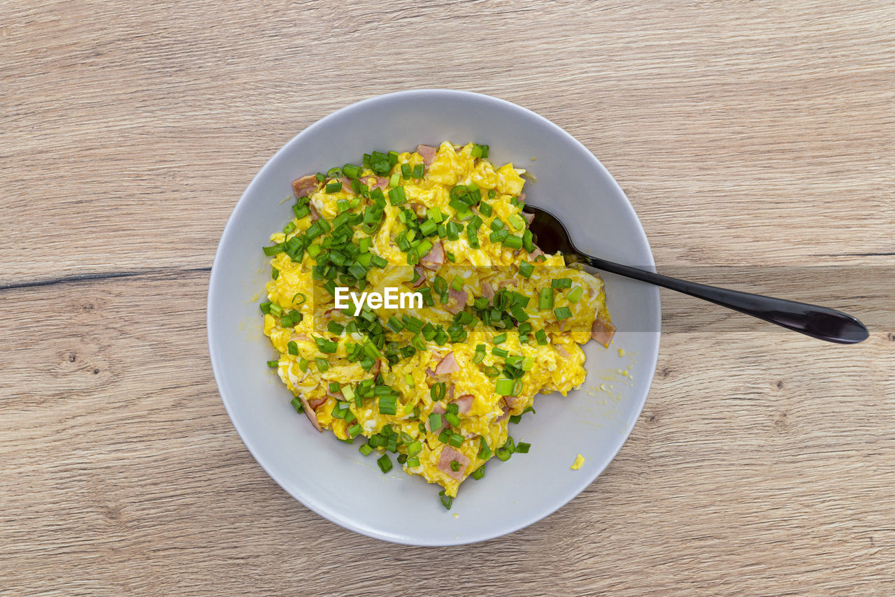Scrambled eggs with chives and pieces of ham, lying on a plate with a fork on a wooden table. 