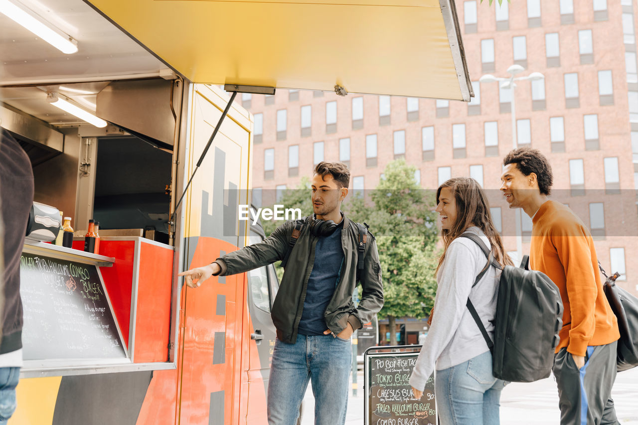 Young man pointing at menu while standing with friends by food truck in city