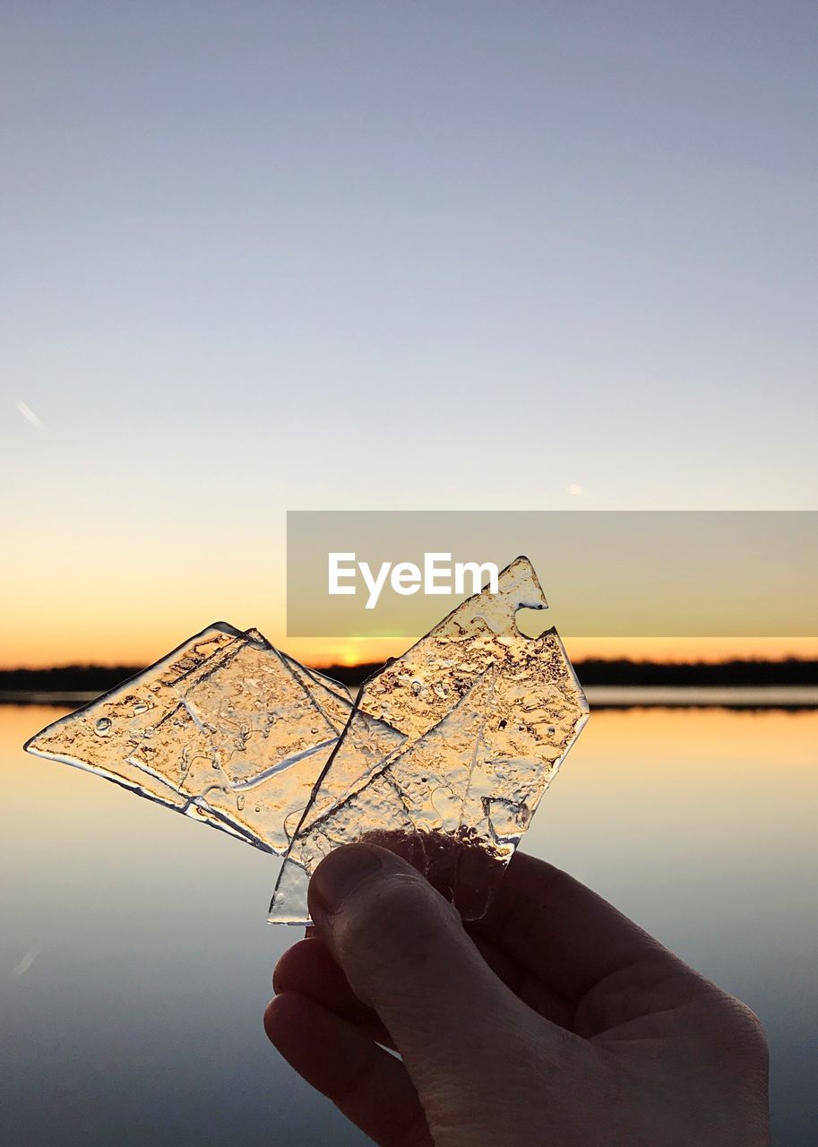 Cropped hand holding ice by lake during sunset