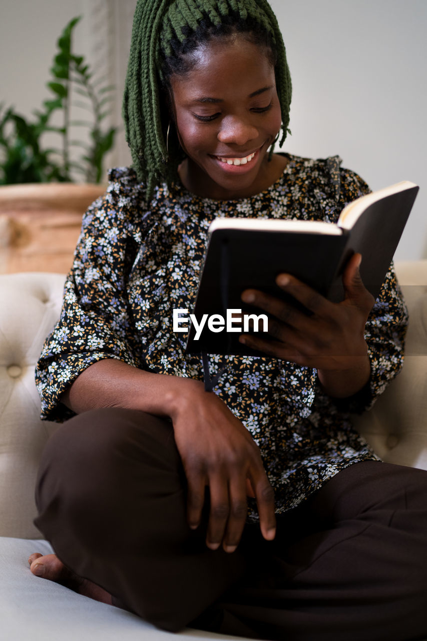 Positive african american female with braids reading interesting book while resting on couch in modern living room at home