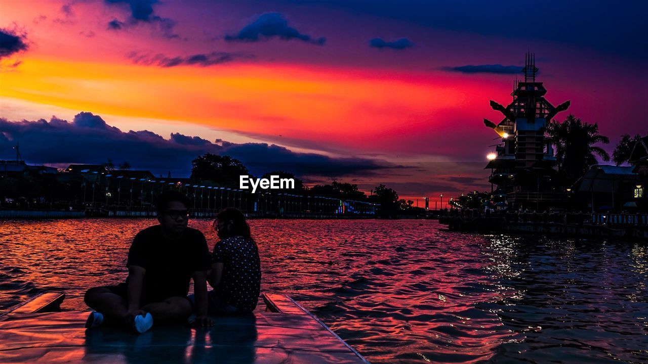 REAR VIEW OF COUPLE SITTING ON WATER AGAINST ORANGE SUNSET SKY