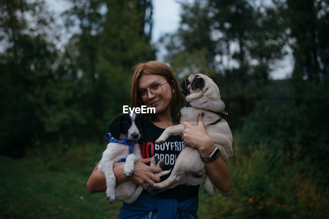 Portrait of smiling woman holding dog standing against trees