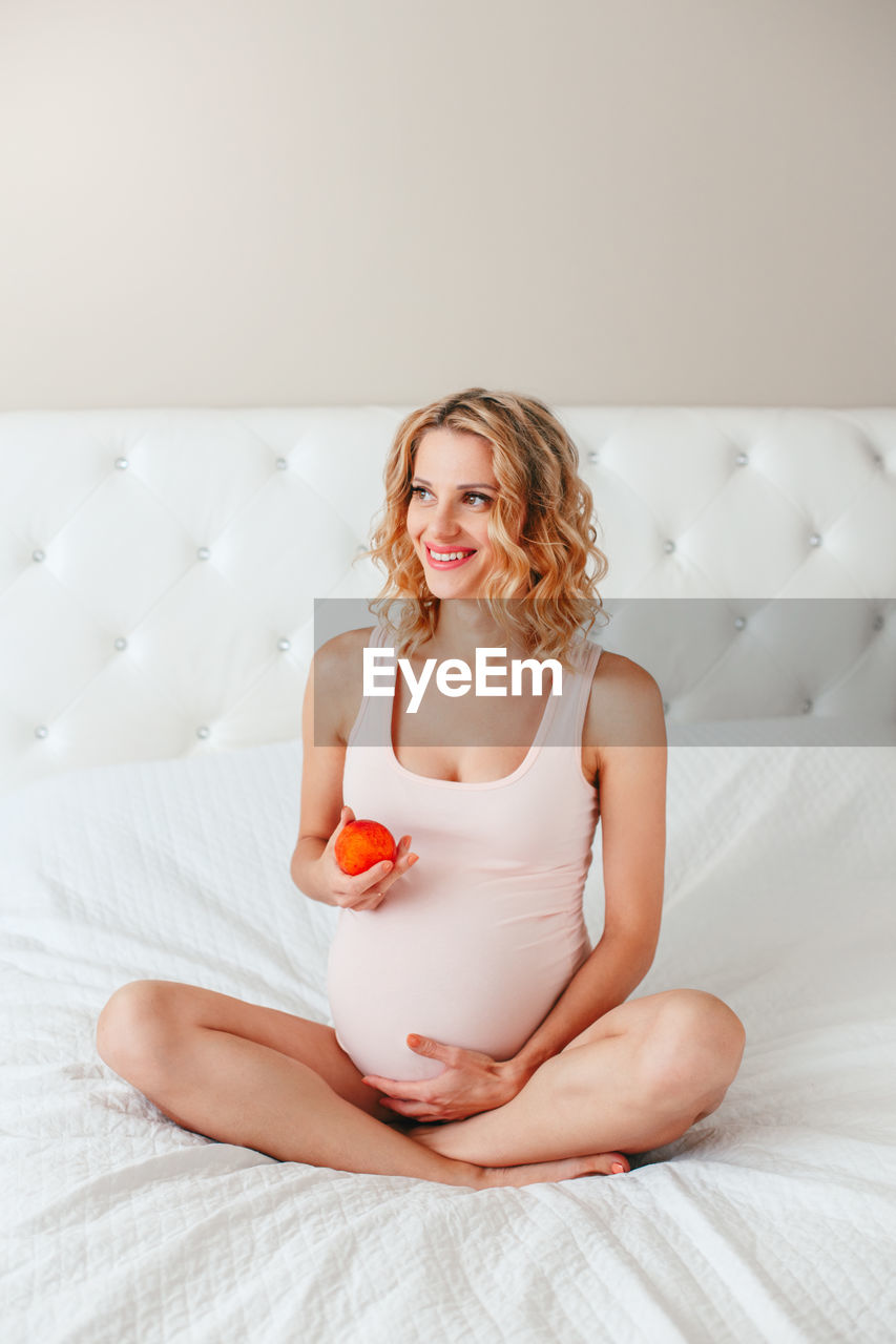 Pregnant smiling woman holding fruit while sitting on bed at home