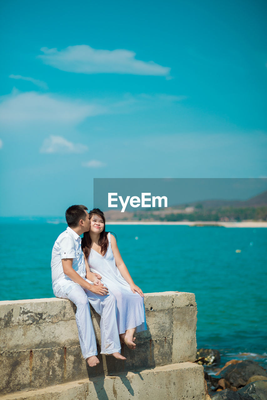 Man kissing woman while sitting against sea