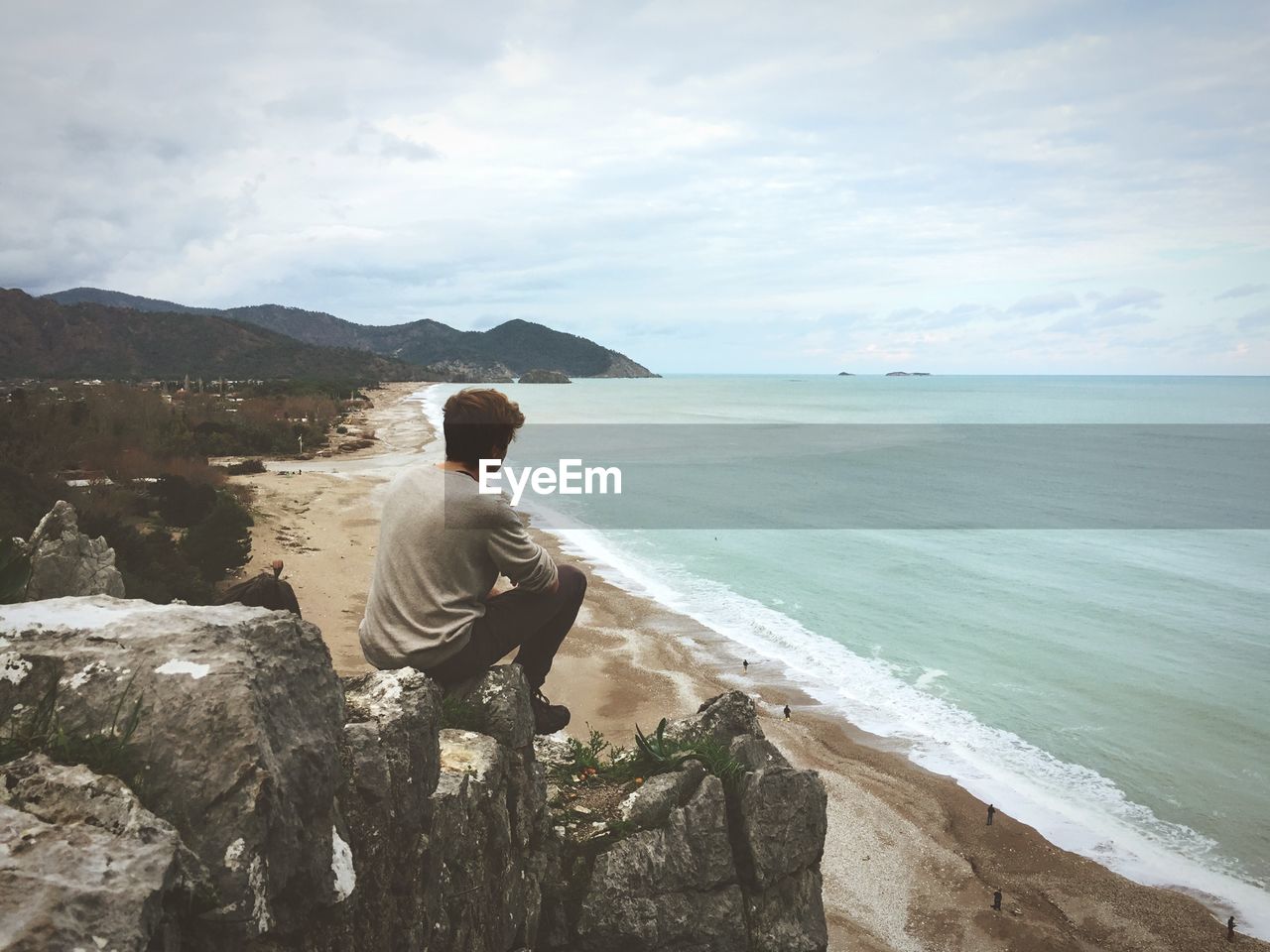 Rear view of man sitting on rock viewing sea against sky