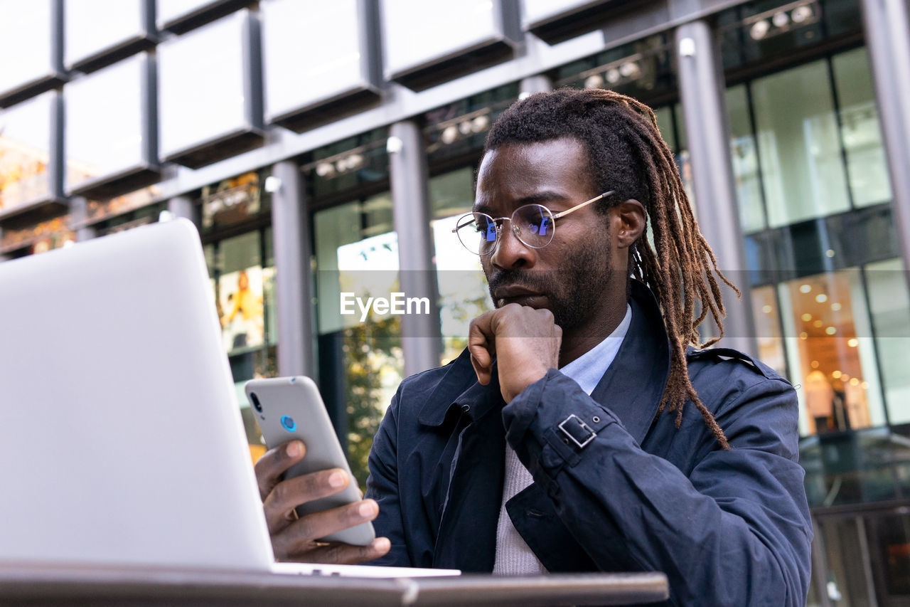 Serene african american young man messaging on smartphone sitting at table with a laptop on terrace of shopping center