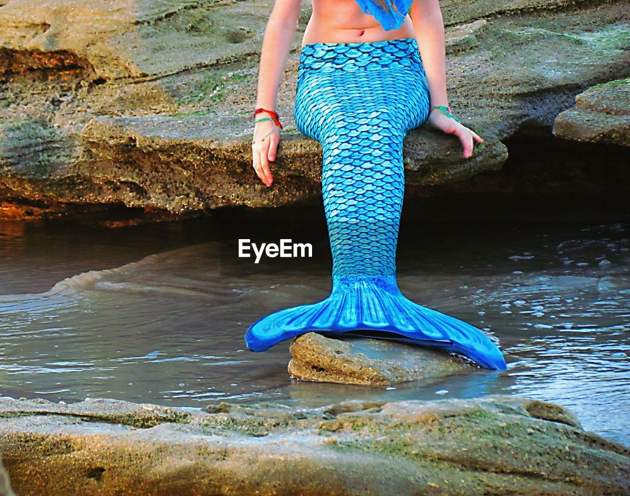Woman wearing blue mermaid dress sitting on rock at beach