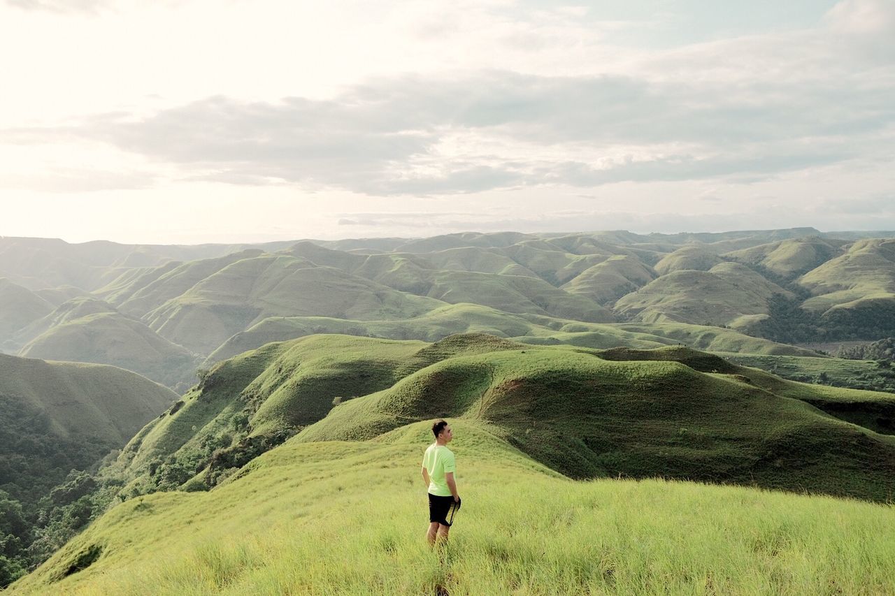 Side view of young man standing on grassy mountain against sky