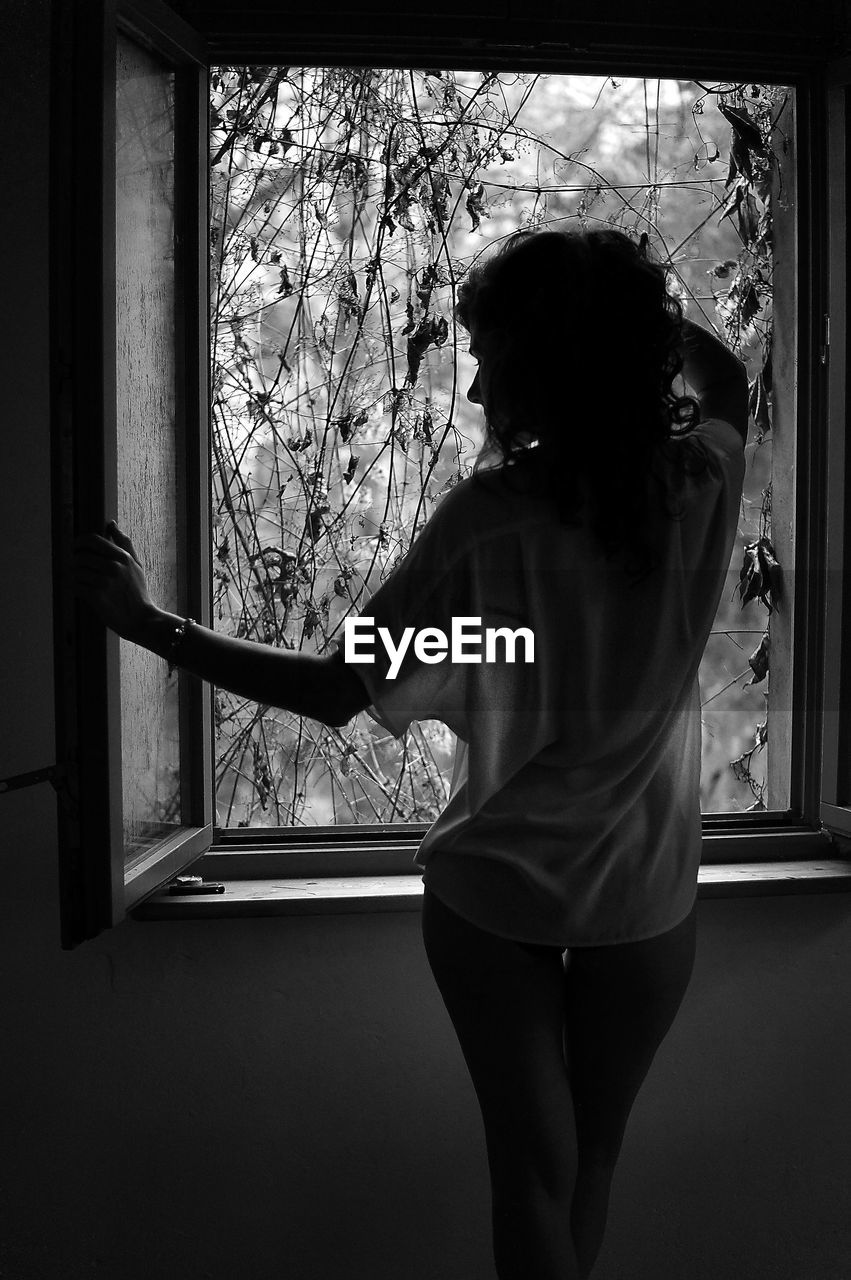 Rear view of sensual woman standing by window at home