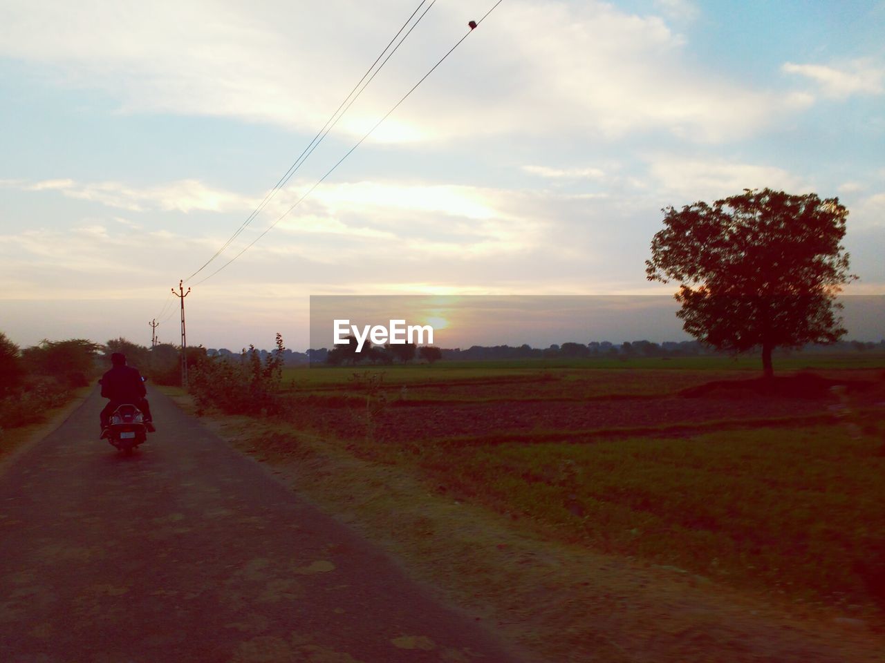 Rear view of person riding scooter on street by grassy field against cloudy sky