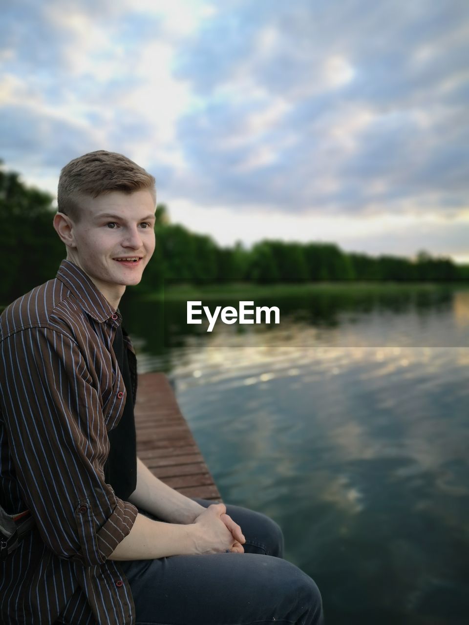 PORTRAIT OF TEENAGE BOY SITTING AGAINST LAKE