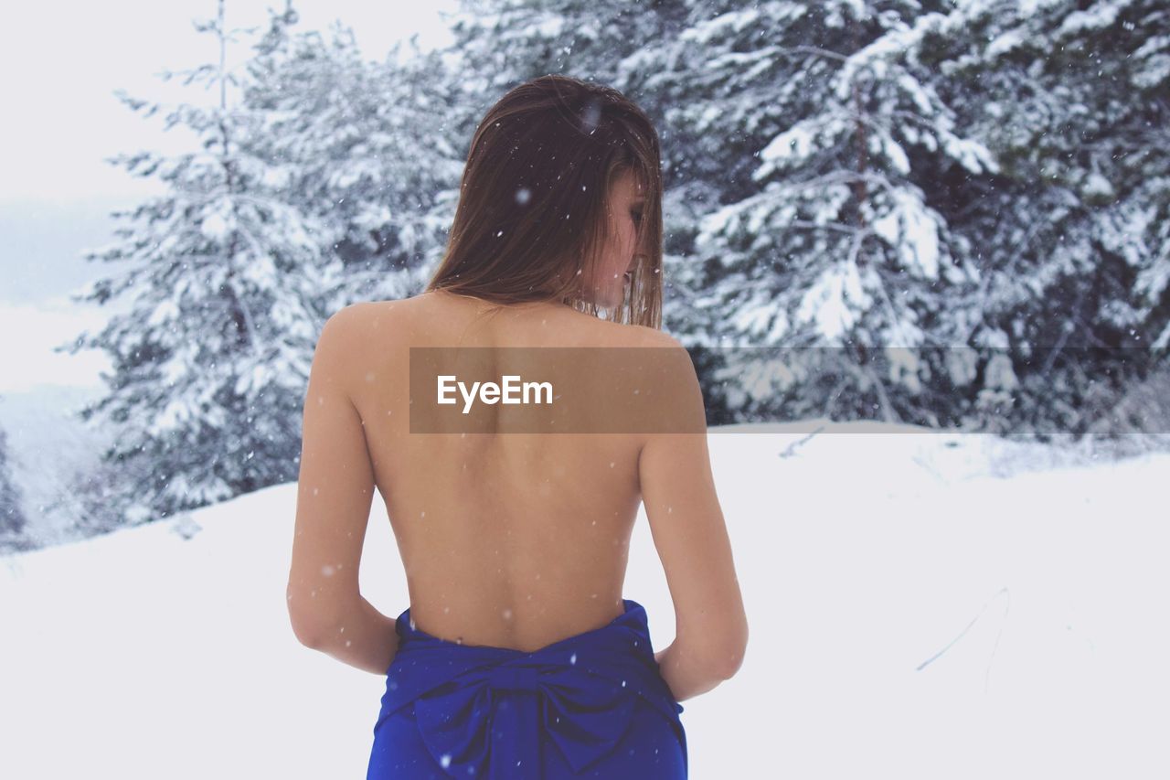 Rear view of shirtless young woman standing on snow against trees