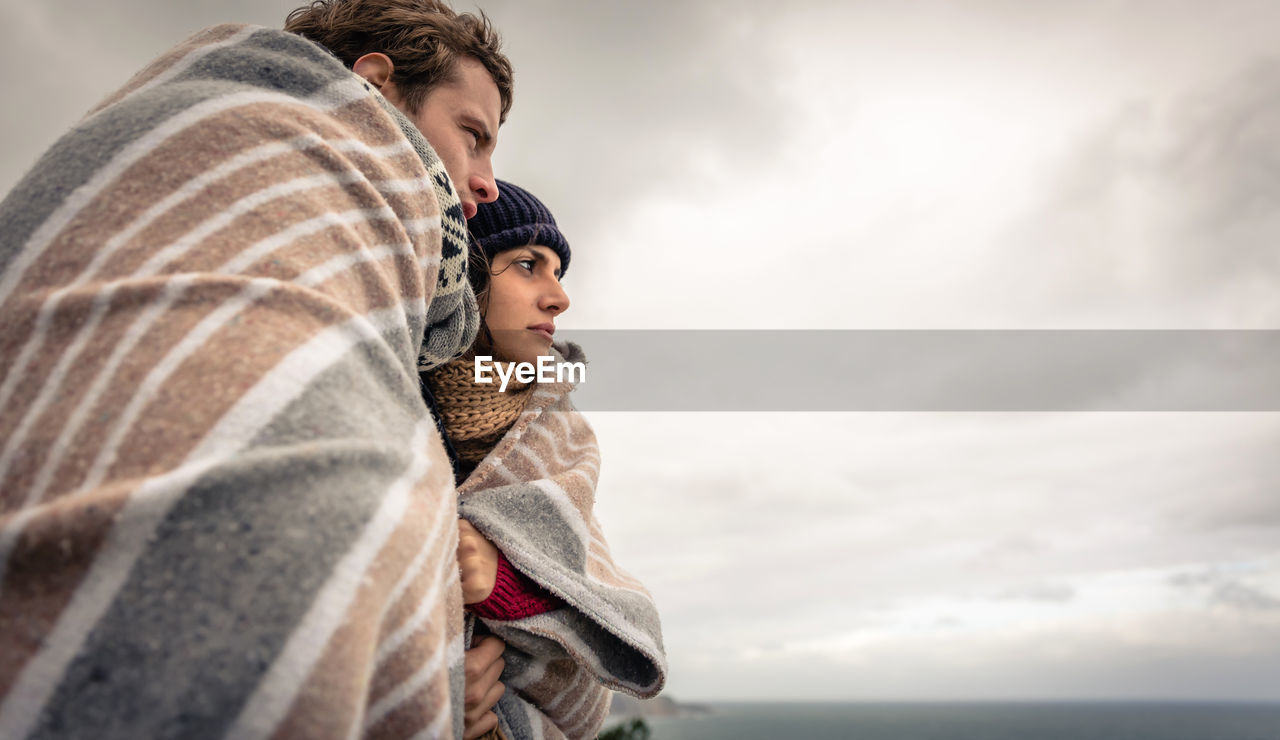 Couple covered with blanket standing against sky