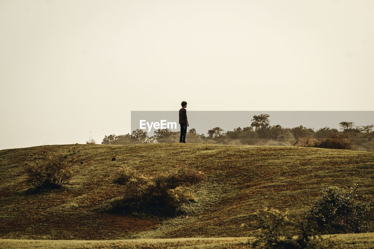 Side view of young man standing on hill against clear sky during sunny day