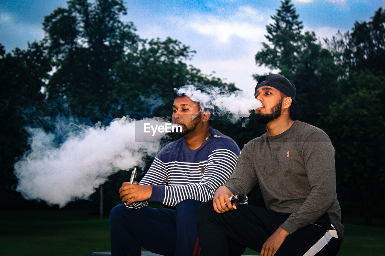Young friends exhaling smoke while sitting at park during sunset