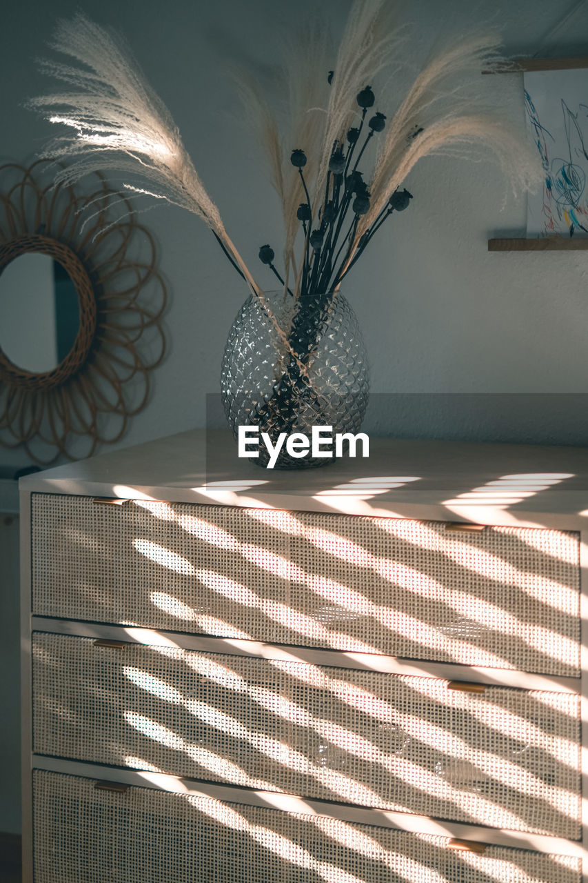 Vase of dry flowers on a wooden dresser enlightened by soft sunlight throwing shadows on the surface