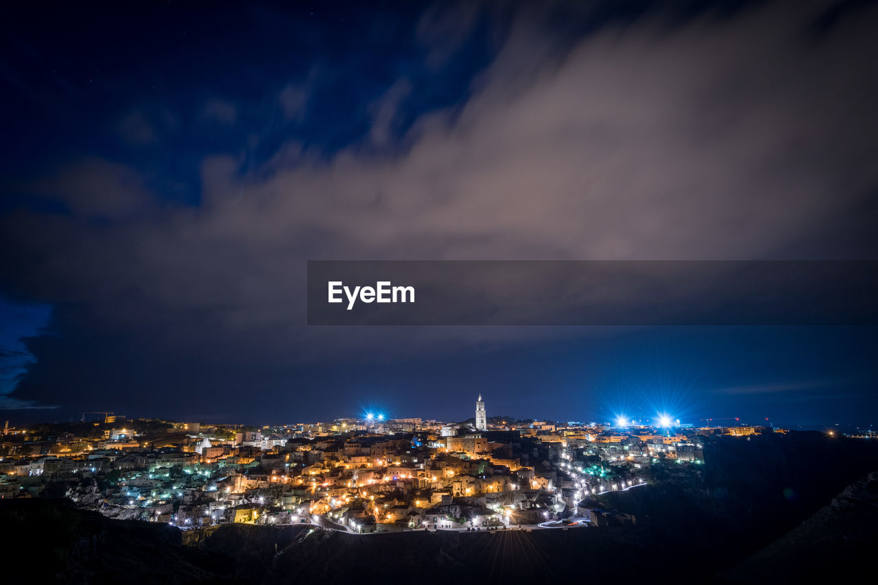 Illuminated cityscape against cloudy sky at night