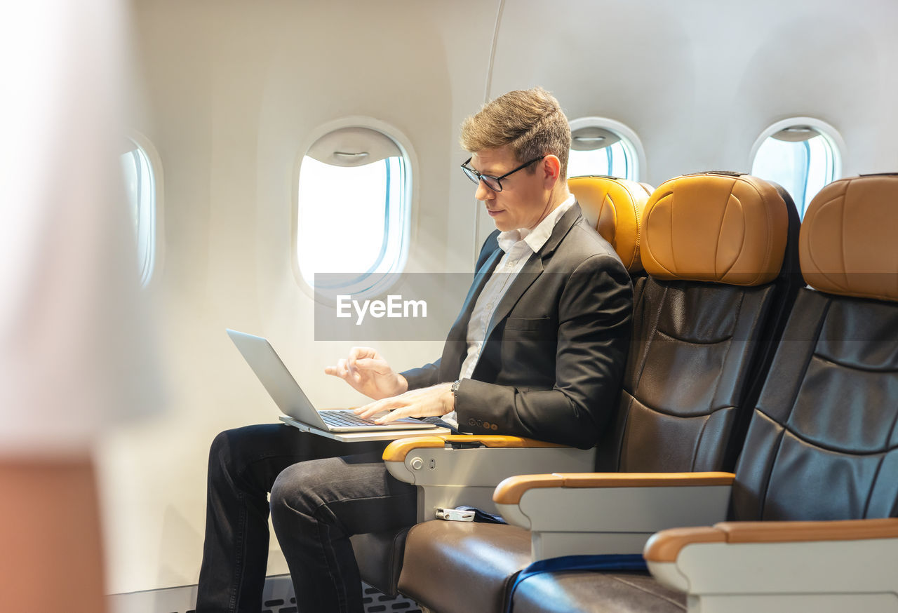 During a flight, a businessman in formal attire and glasses looks out the plane window 