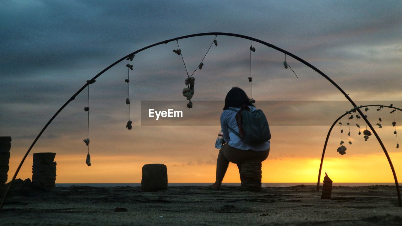 REAR VIEW OF MAN STANDING ON SHORE AGAINST SKY DURING SUNSET