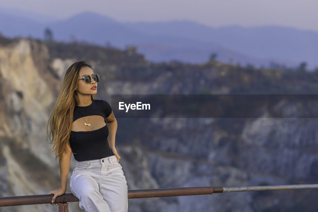 Woman wearing sunglasses standing on railing against mountain