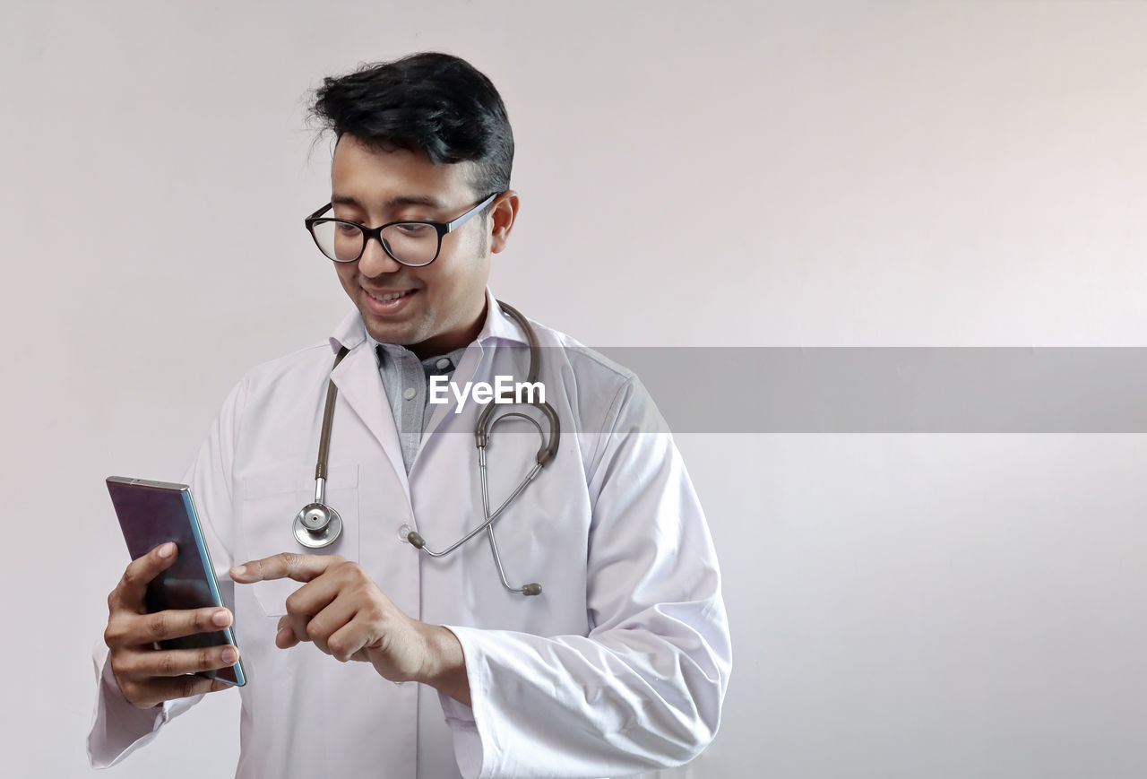 Smiling doctor using mobile phone against wall