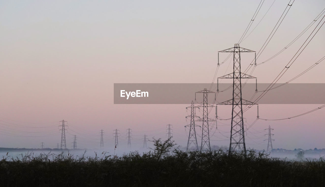 Low angle view of electricity pylons on field against sky during sunrise