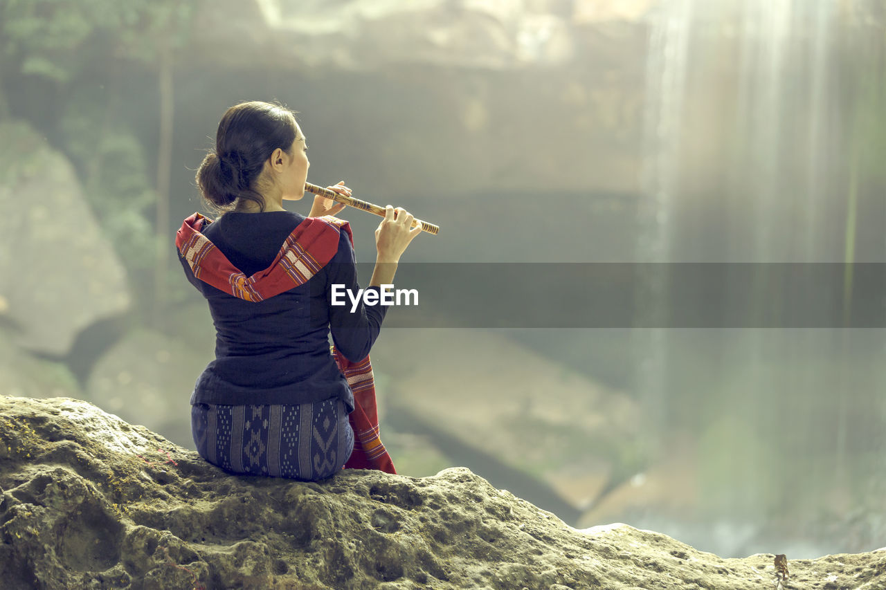 Rear view of woman playing flute while sitting on rock against waterfall