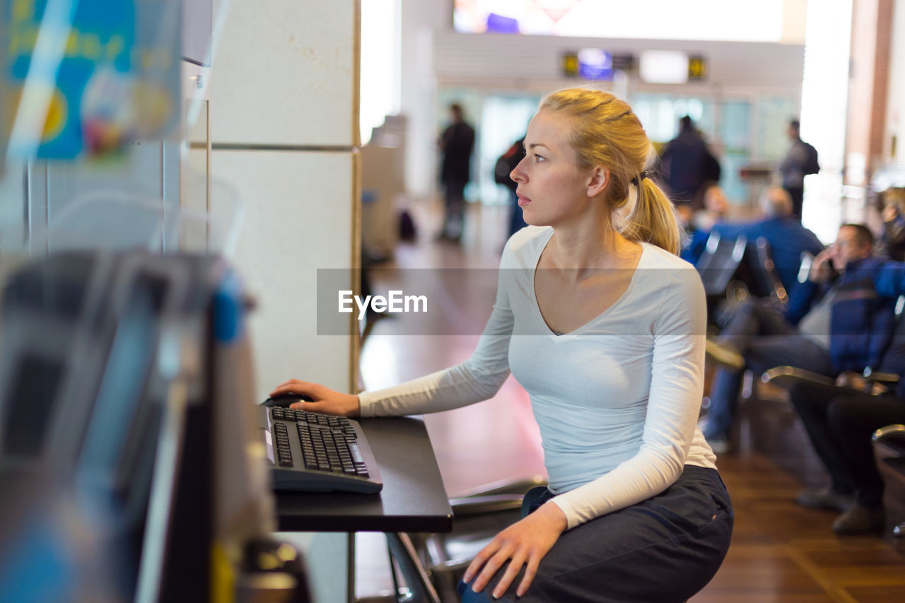 YOUNG WOMAN USING PHONE WHILE SITTING ON LAPTOP IN OFFICE