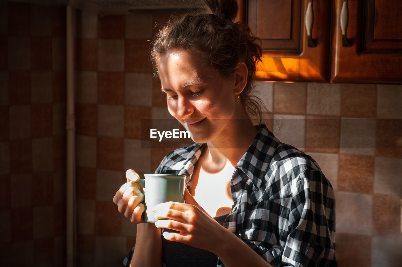 Young woman hold and enjoy aromatic cup of coffee in morning sunlight at home