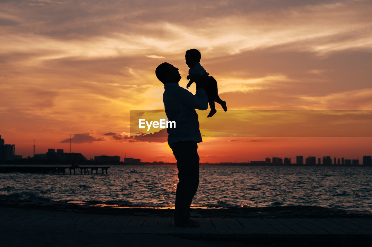 Silhouette man carrying son while standing by sea against sky during sunset