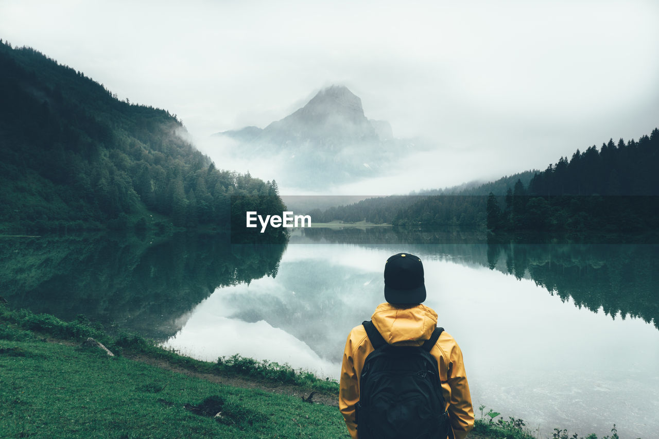 Rear view of man looking at obersee lake by mountains in foggy weather