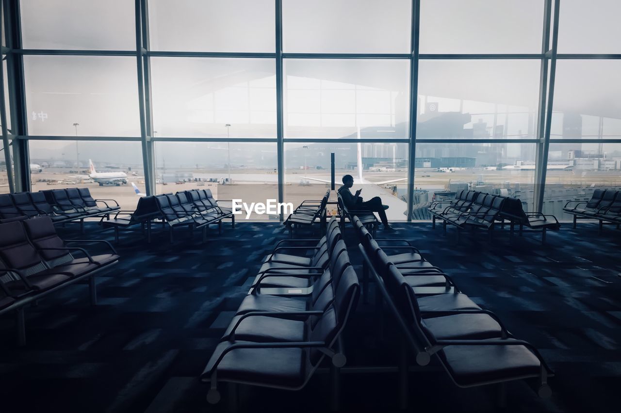 PEOPLE SITTING ON CHAIRS AT AIRPORT AGAINST GLASS WINDOW