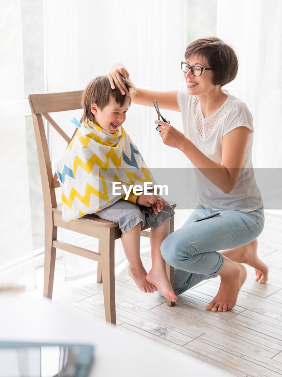 Mother cuts her son's hair by herself. little boy sits, and holds pair of scissors. 