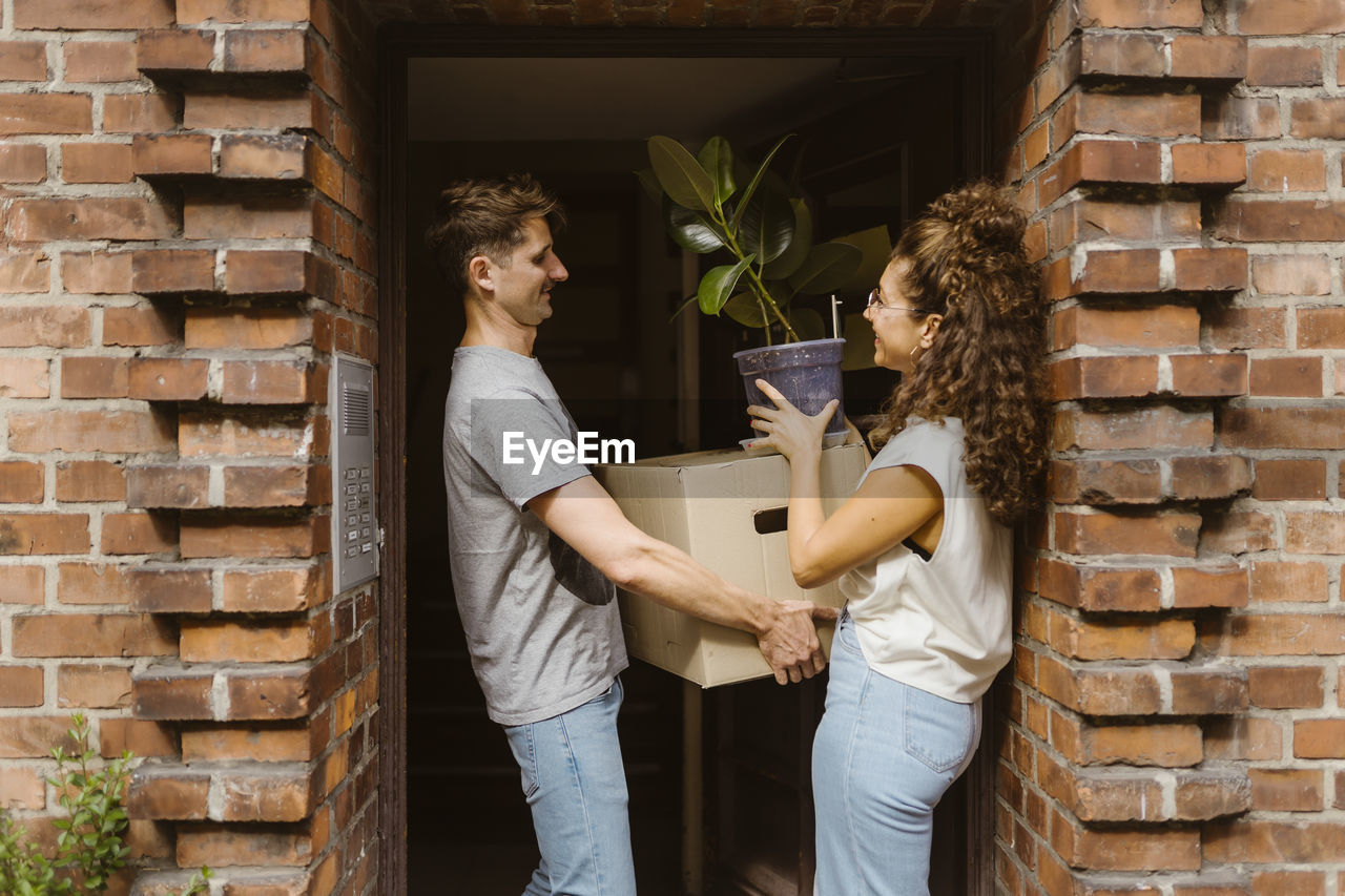 Happy couple with cardboard box and potted plant talking to each other at doorway