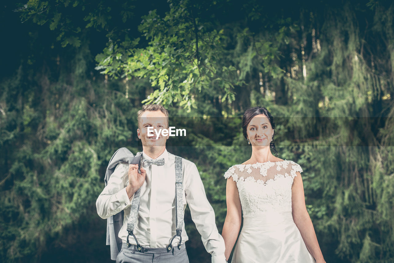Portrait of smiling bride and groom standing against trees