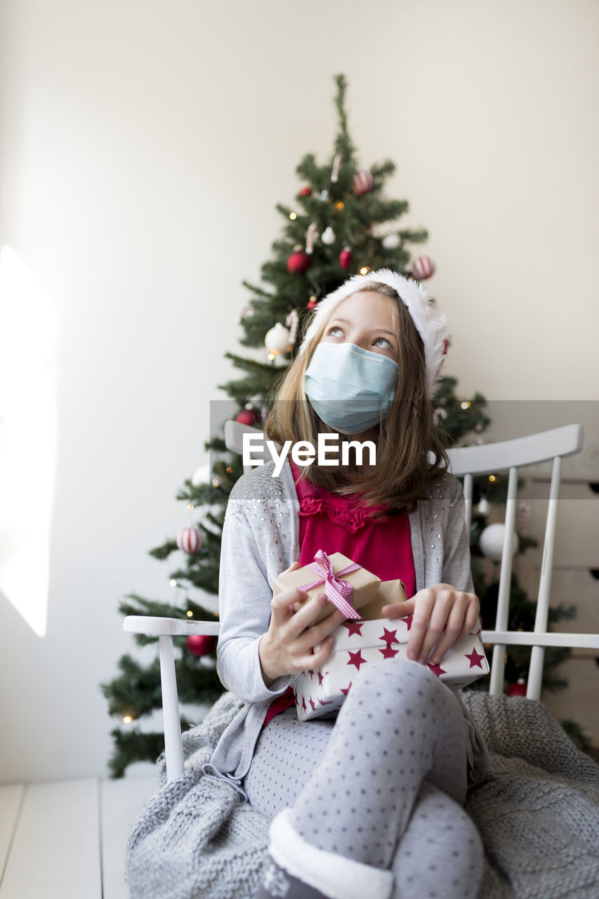 Girl wearing mask holding gifts against christmas tree at home