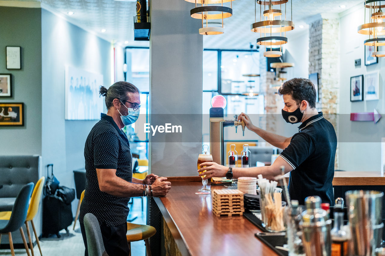 Side view of male customer in medical mask standing at counter and waiting for glass of beer served by barman