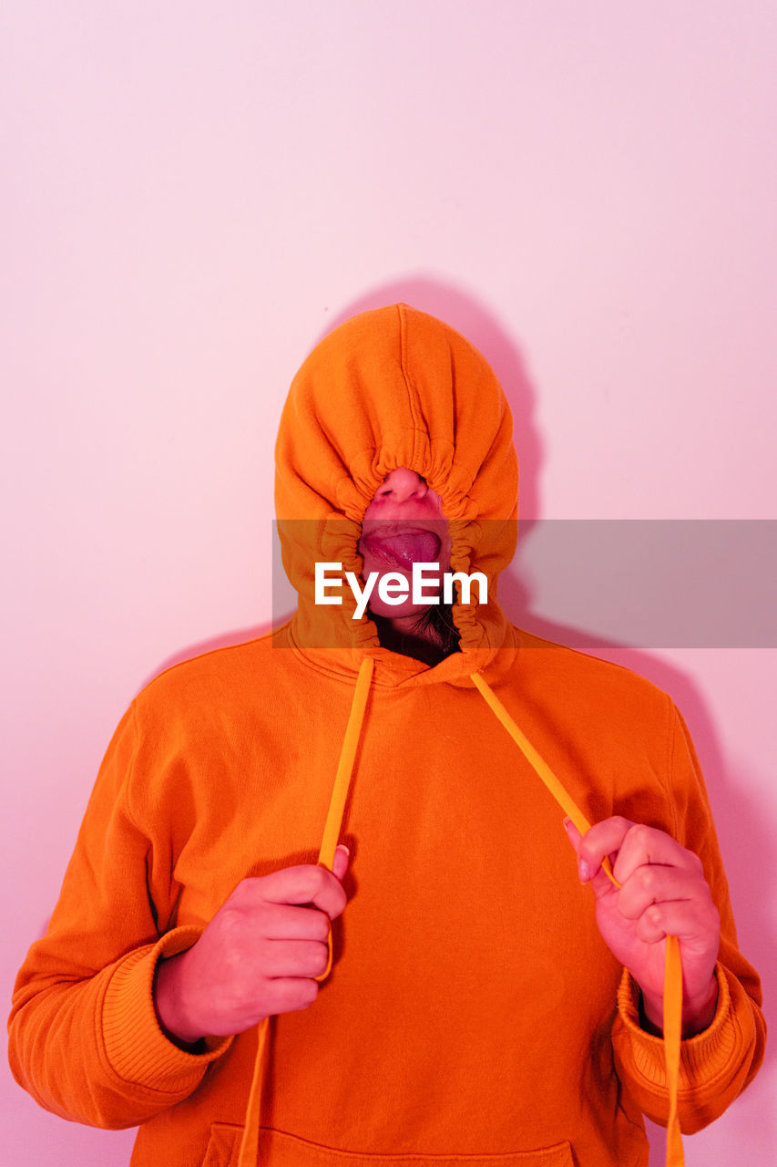 Woman wearing orange hood standing against pink wall