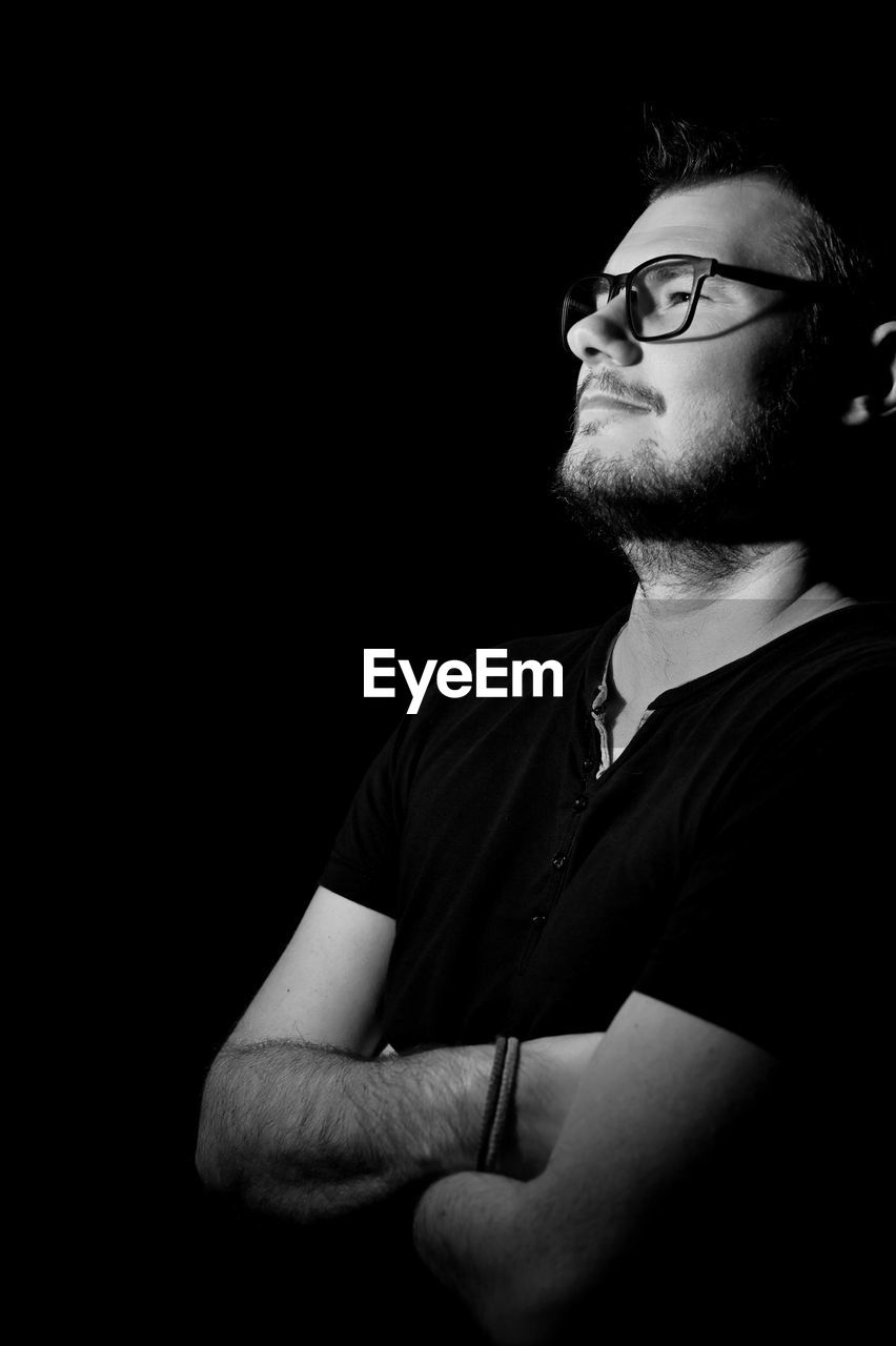 black, black and white, one person, glasses, eyeglasses, monochrome photography, adult, black background, darkness, monochrome, studio shot, men, portrait, indoors, beard, facial hair, white, young adult, copy space, looking, waist up, casual clothing, person, looking away, contemplation, eyewear, fashion, arms crossed, headshot, dark, lifestyles, serious, human face