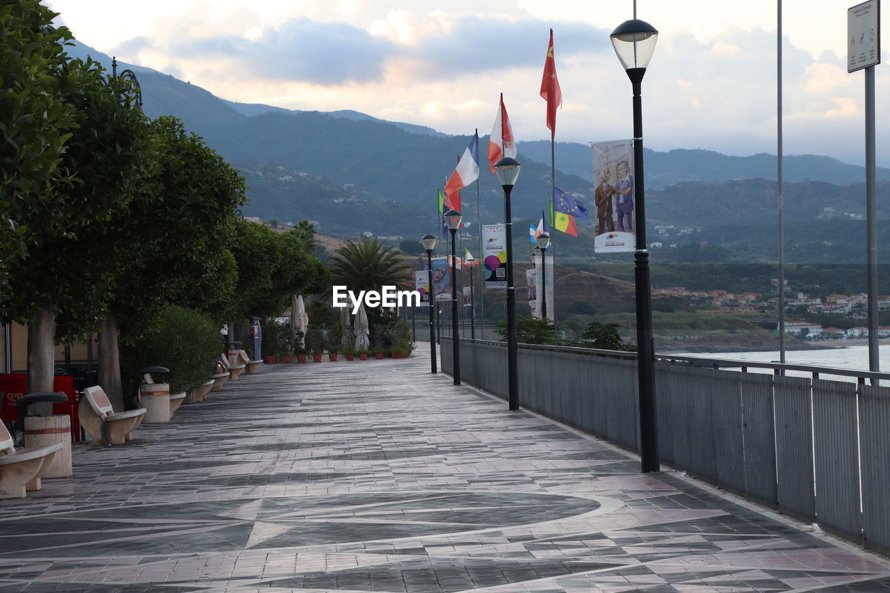 walkway, architecture, mountain, sky, nature, tree, flag, built structure, city, plant, travel destinations, street, cloud, building exterior, street light, boardwalk, no people, water, town, mountain range, outdoors, environment, scenics - nature, travel, building, landscape, patriotism, day, beauty in nature, footpath, tourism, transportation, lighting equipment, tranquility, road, vacation