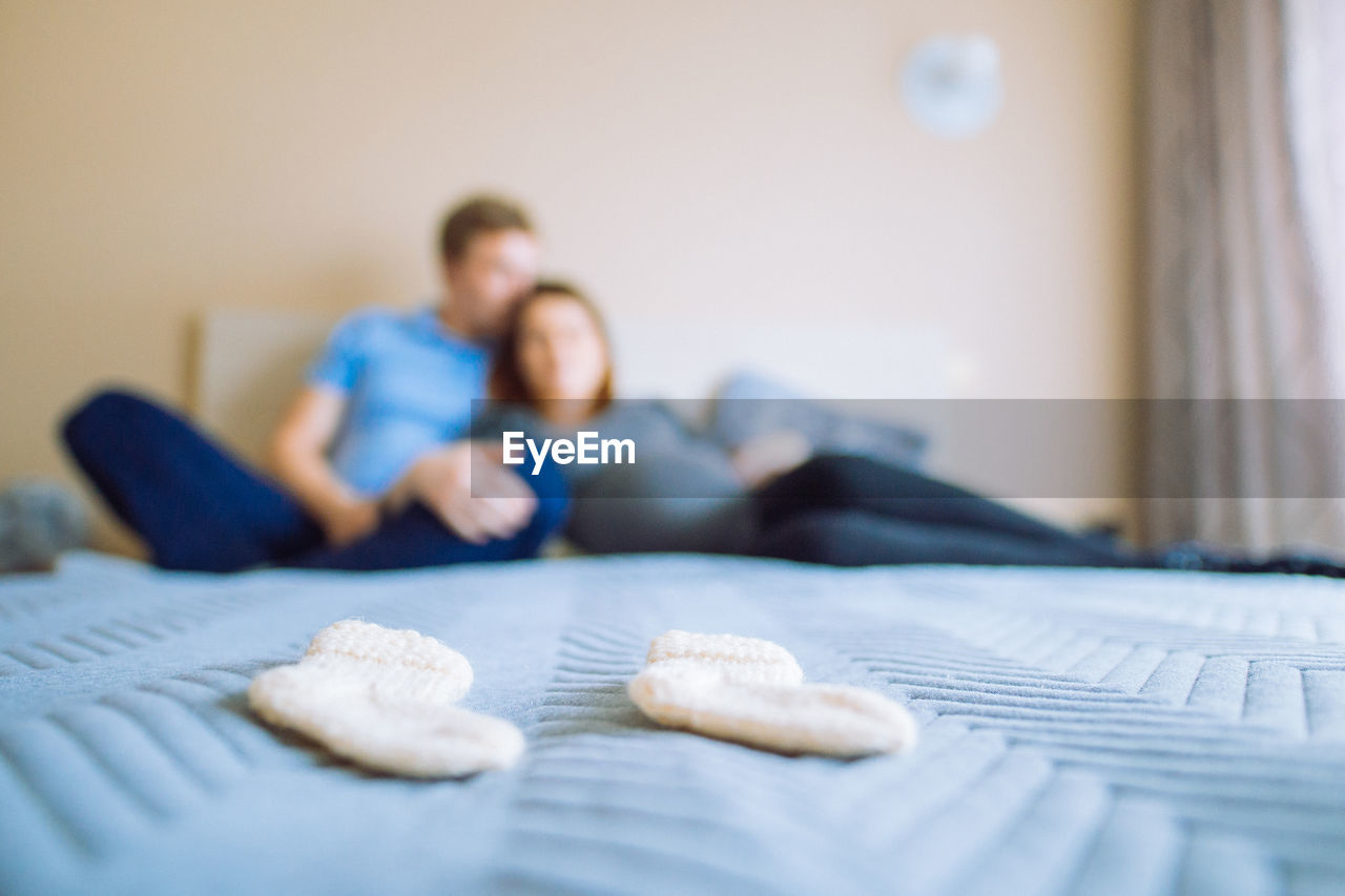 Close-up of baby socks against man with pregnant woman relaxing on bed at home