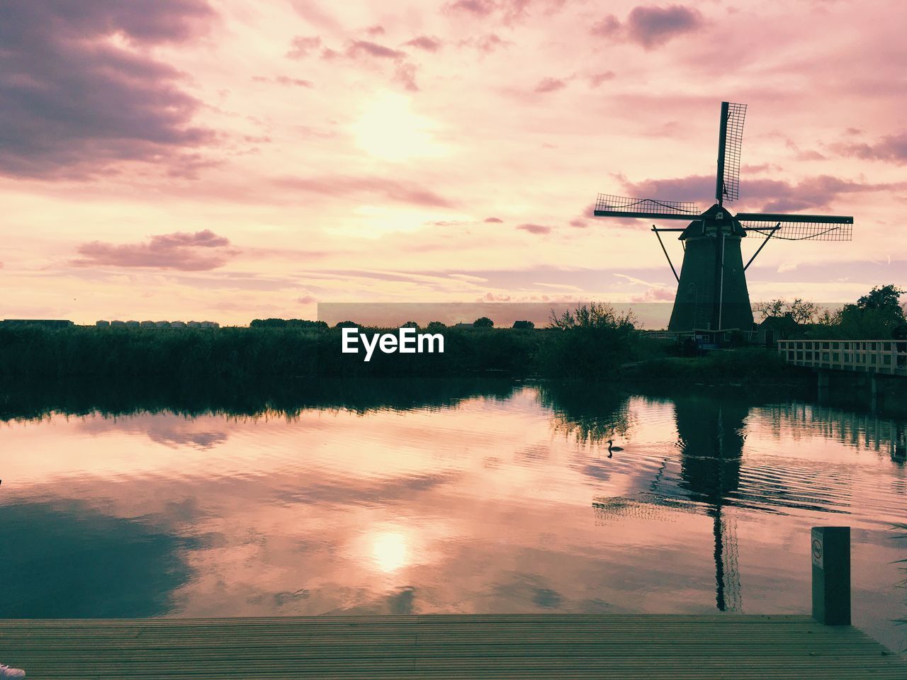 Traditional windmill by lake against cloudy sky during sunset