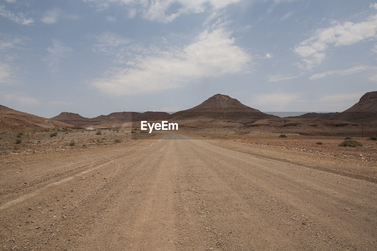 Empty dirt road towards mountains against sky