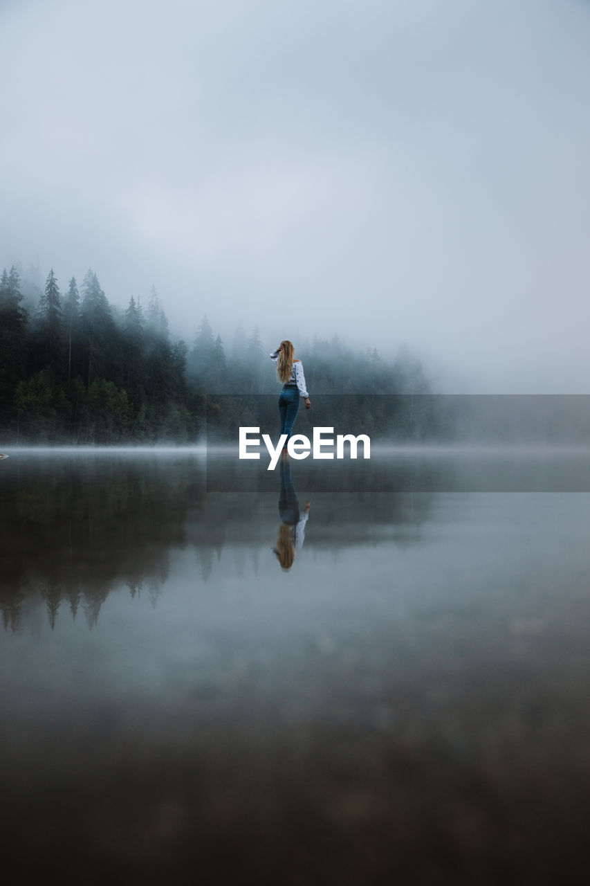 Young caucasian blonde woman standing in the calm forest foggy lake.	