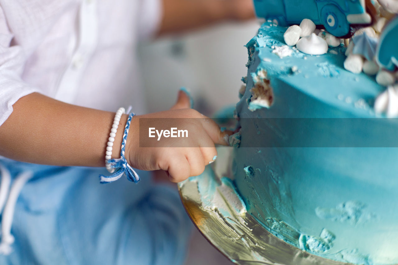 Baby boy in blue pants and shirt standing on the floor next to the cake in blue and white balloons